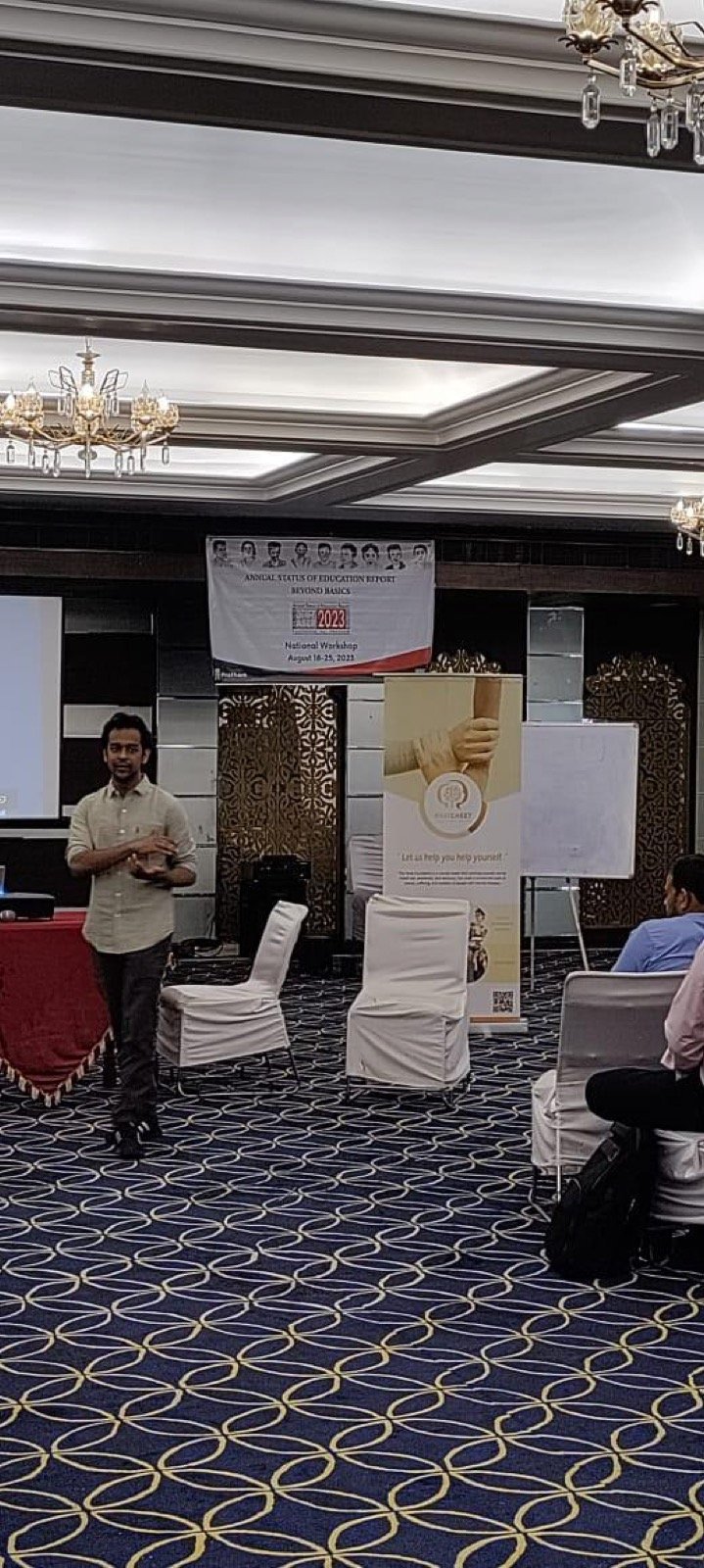 A speaker addresses the room from the front of a hotel conference hall, beneath a banner reading ASER 2023 Beyond Basics National Workshop, with a Baatcheet standee alongside.