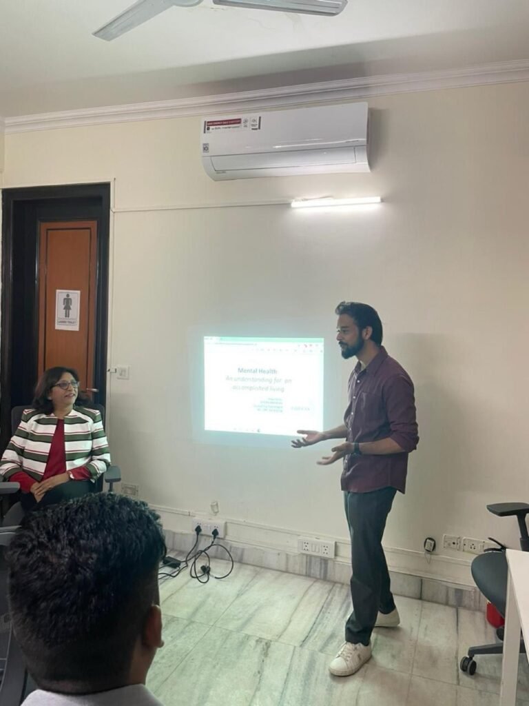 A speaker presents a slide headed Mental Health to a small seated audience in a meeting room.