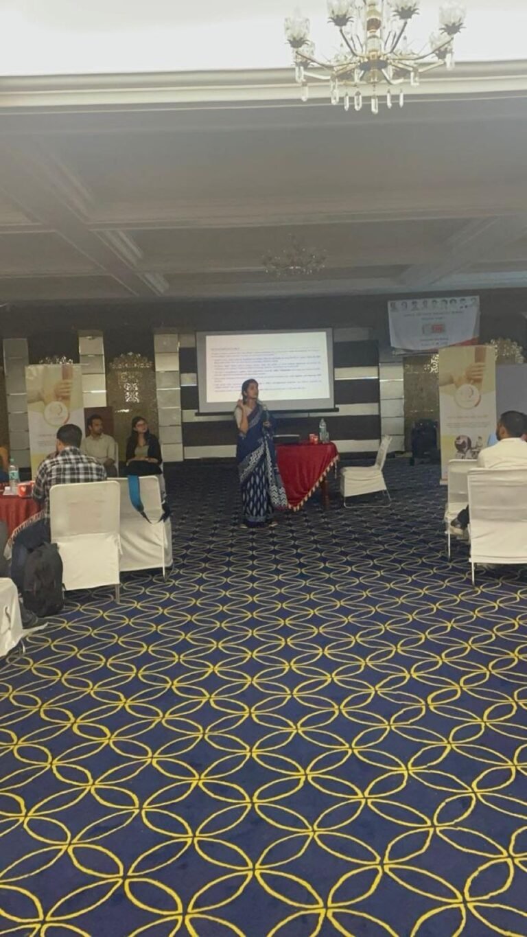 A presenter speaks beside a projected slide in the conference hall while attendees follow from tables around the room.
