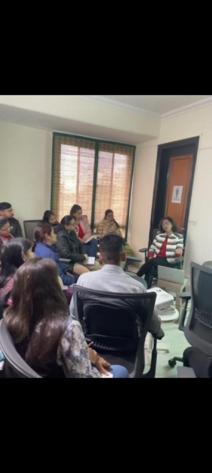 A group sits in a loose circle in a small room, listening to a facilitator seated by the window.