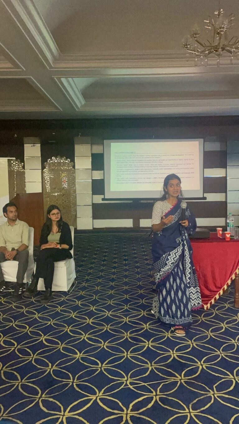A presenter in a sari speaks to the conference hall while two colleagues listen from chairs at the front.
