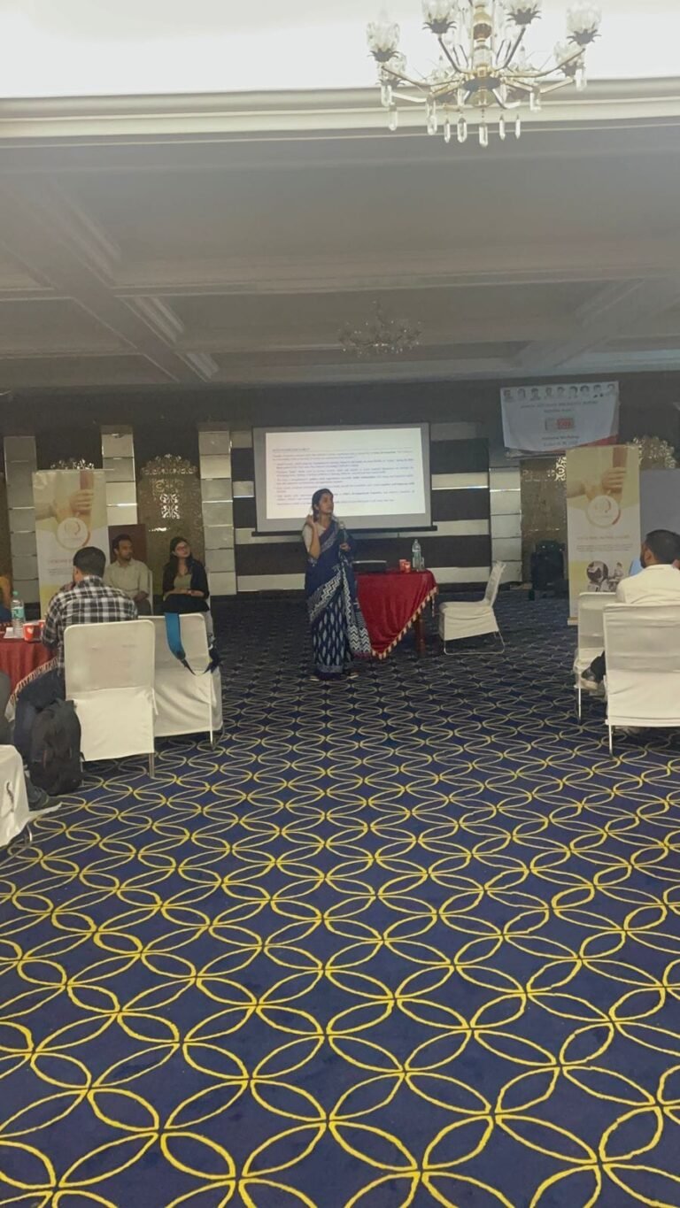 A presenter speaks from the centre of the conference hall, with a projected slide and a draped table behind her.